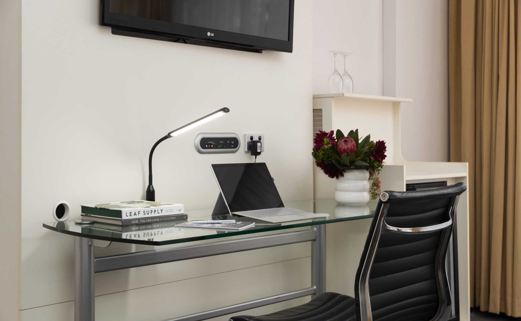 A modern desk setup with a laptop, a book, a desk lamp, and a decorative flower arrangement next to a television.