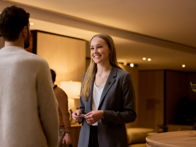 A smiling woman in a gray blazer engages in conversation with a person in a light sweater, set in a modern interior.