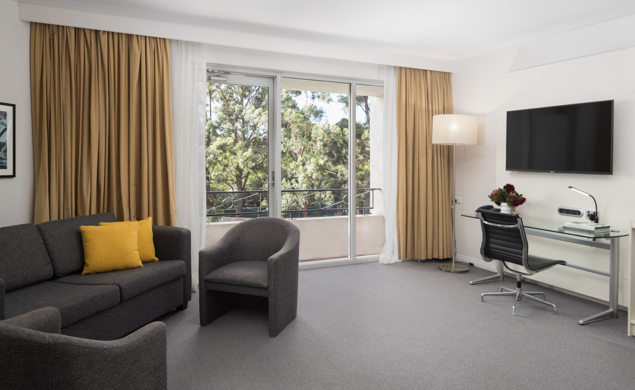 A modern hotel room featuring a gray sofa with a yellow pillow, armchairs, desk with a chair, and large windows overlooking trees.