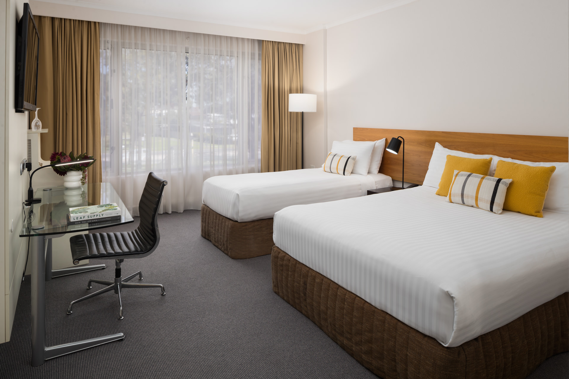 A hotel room featuring two beds with white linens, a desk with a chair, and large windows with sheer curtains.